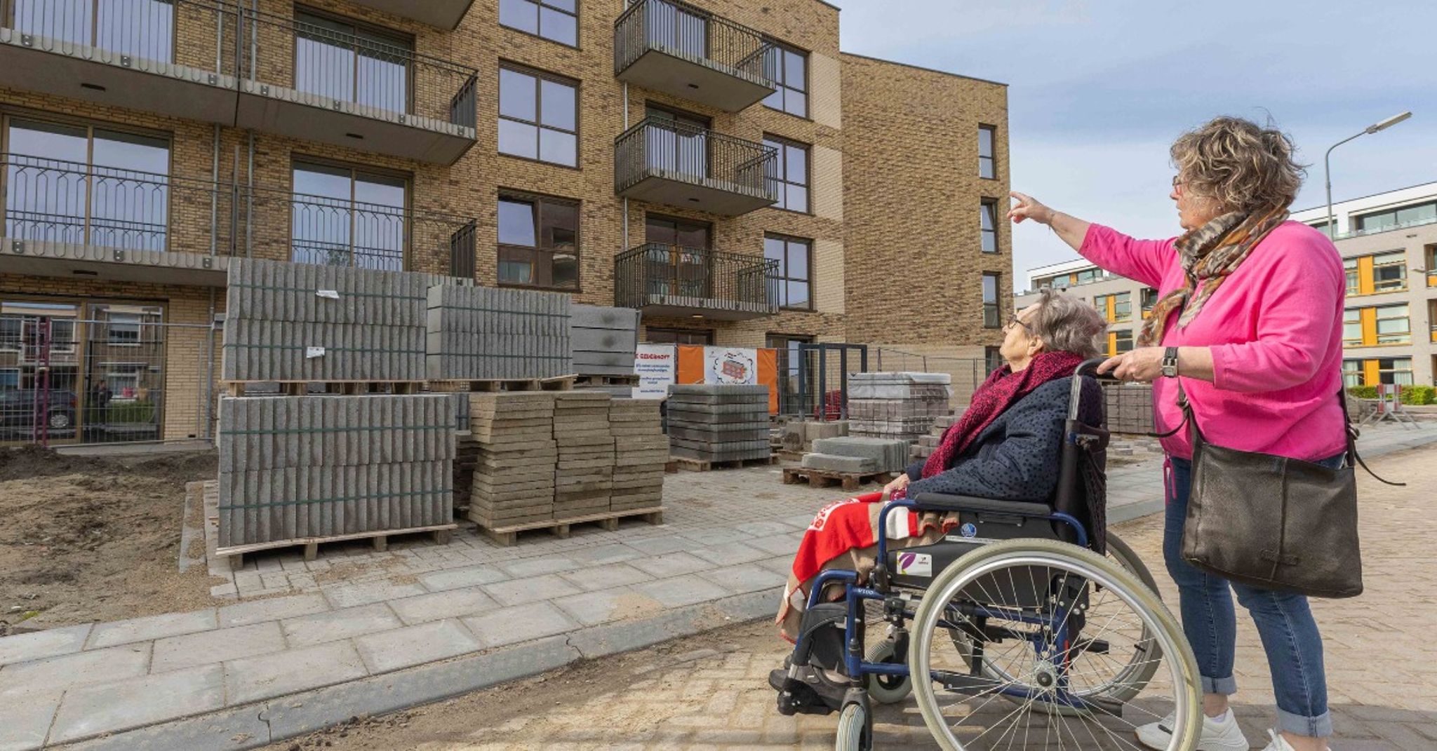 Een bouwplaats voor een meerlaagse bakstenen flat met balkons. Op de voorgrond zit een oudere persoon in een rolstoel met een rode deken, naast een staande persoon in roze jas en blauwe jeans die naar het gebouw wijst. Er liggen stapels bouwmaterialen op pallets, zoals stenen en tegels. Op de achtergrond zijn moderne gebouwen met grote ramen zichtbaar.
