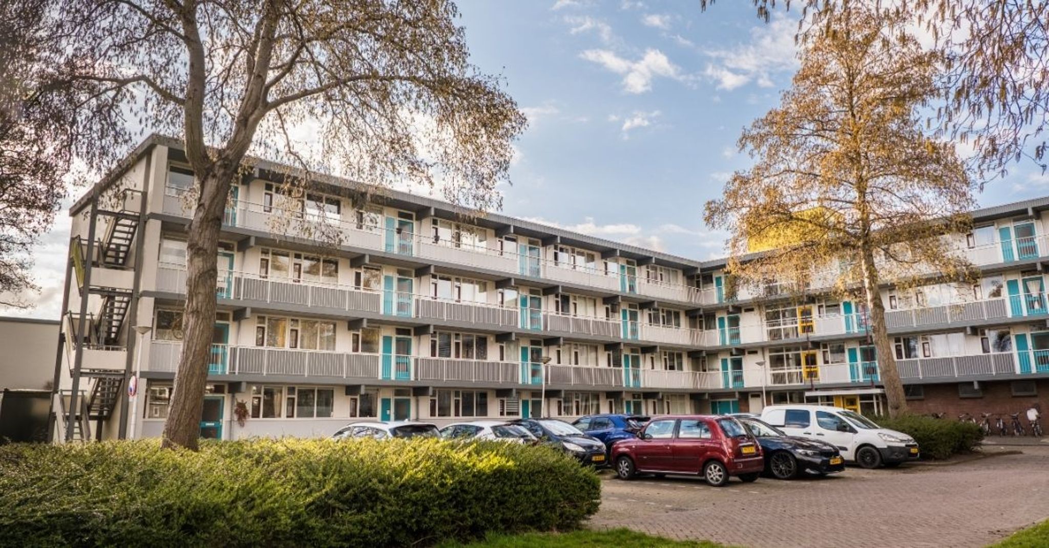 Een vier verdiepingen tellend woongebouw met witte en grijze gevels en turquoise accenten op de deuren. Het gebouw heeft meerdere balkons en ramen. Op de voorgrond staan bomen en zijn auto's geparkeerd. De lucht is deels bewolkt, wat wijst op een daglichtsituatie.