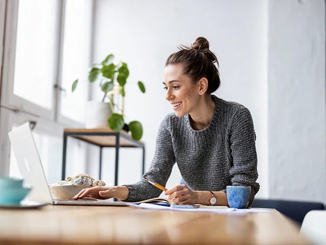 Een jonge lachende vrouw met grijze truin achter een laptop in een witte woning met plant.