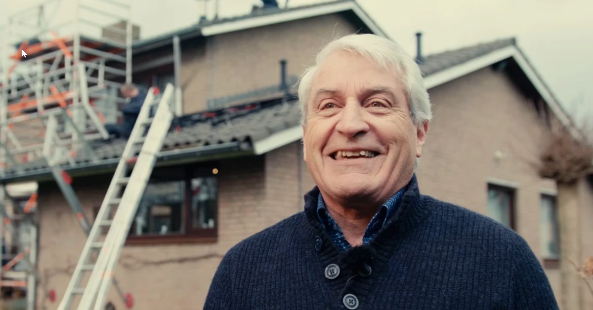 Lachende oudere man voor een woning waar installateurs zonnepanelen op aan het leggen zijn.