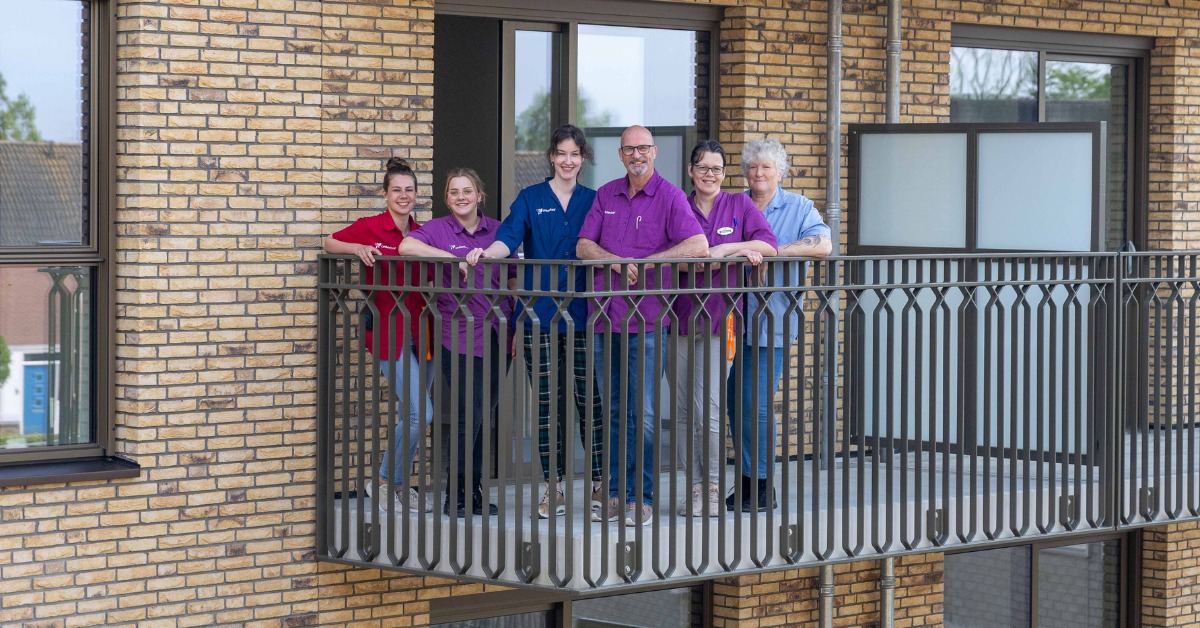 Zes zorgmedewerkers staan op een balkon van een bakstenen gebouw en leunen tegen de railing. Ze dragen shirts in verschillende kleuren, waaronder rood, blauw en paars. 