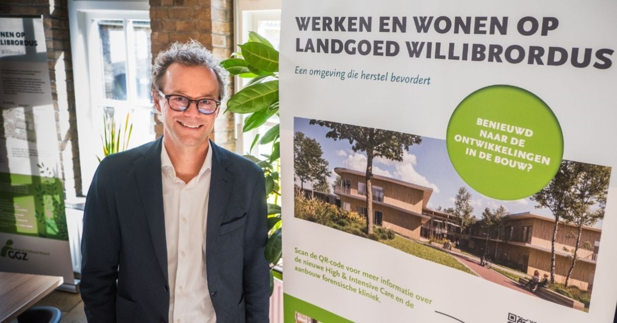 Daan de Jong, directeur bedrijfsvoering van GGZ NHN, naast een groot bord met daarop informatie over een nieuw gebouw.