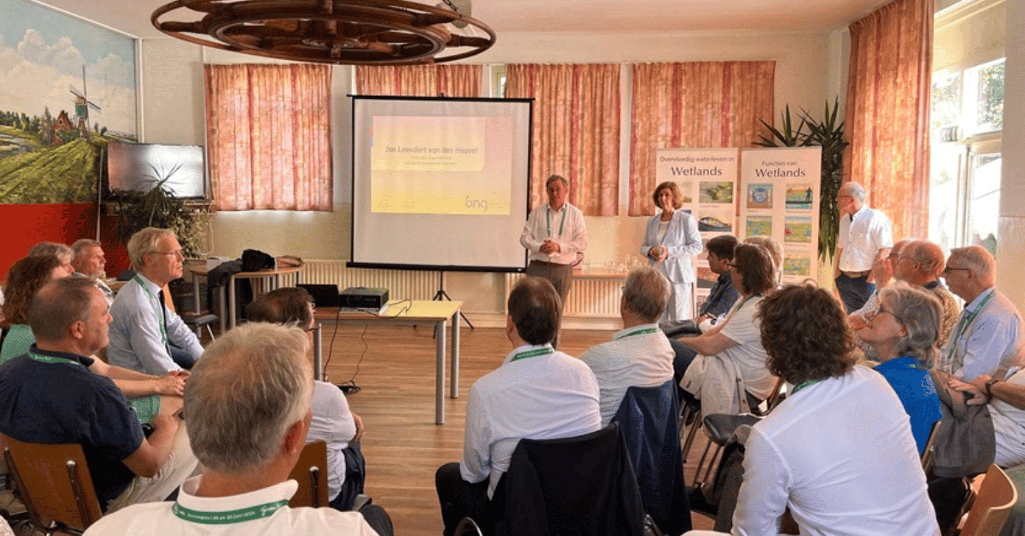 Twee mensen geven een presentatie in een kleine zaal. De aanwezigen zitten om hen heen.